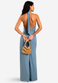Combinaison bleu ciel avec dos nu, lien derrière le cou, jambes larges, tissu texturé. Tient un petit sac à main frangé de ton doré.