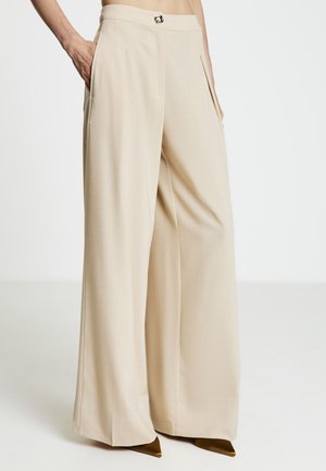 Pantalon large beige avec plis à l'avant, poches latérales et une boucle métallique décorative à la taille. Texture de tissu lisse.
