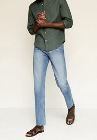 Camisa verde de manga larga con botones y bolsillo en el pecho; jeans azul claro de corte recto; sandalias marrones con tiras.