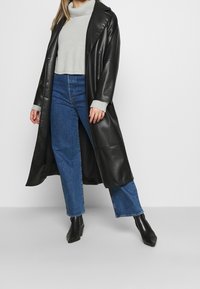 Svart läder lång kappa över en grå stickad tröja och blå vida jeans, kombinerat med svarta spetsiga ankelstövlar. Slät textur med en figursydd silhuett.