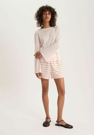 Femme debout portant un haut à manches longues rose clair, un short blanc avec de fines rayures roses, et des sandales tressées noires sur fond uni.