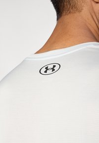 Camiseta deportiva blanca con un cuello redondeado y un logo negro estampado en la parte trasera. El material parece suave y de textura ligera.