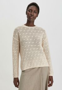 Beige gestrickter Pullover mit einem strukturierten, offenen Muster und langen Ärmeln. Rundhalsausschnitt und lässige Passform. Kombiniert mit einem hellen Rock.