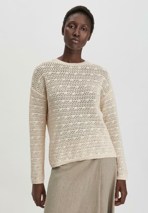 Beige gebreide trui met een gestructureerd, open geweven patroon en lange mouwen. Ronde halslijn en een relaxte pasvorm. Gecombineerd met een lichtgekleurde rok.