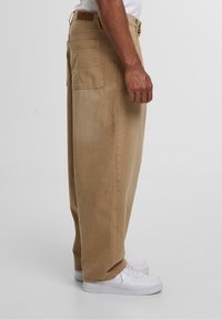Beige brede bukser lavet af bomuld, med sidelommer og en baglomme, parret med hvide sneakers.