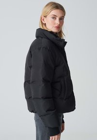 Giacca a vento nera con colletto alto, dotata di chiusura con zip frontale e tasche laterali. Il design è squadrato, con una finitura opaca e polsini a costine.