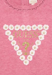 Maglione rosa con un design triangolare arricchito da fiori bianchi e testo glitterato dorato con la scritta "GUESS?" accentuato da ulteriore testo.