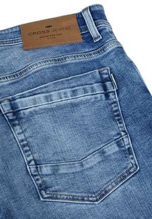 Achterzak en riemlus van middelblauwe spijkerbroek met een bruine leren patch met de tekst "CROSS JEANS Denim for Life."