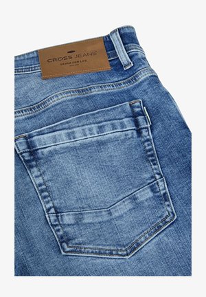 Achterzak en riemlus van middelblauwe spijkerbroek met een bruine leren patch met de tekst "CROSS JEANS Denim for Life."