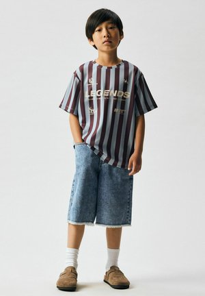 Chłopiec stojący z jedną ręką w kieszeni, ubrany w pasiastą koszulkę z napisem "LEGENDS", dżinsowe krótkie spodenki, białe skarpetki i brązowe wsuwane buty.