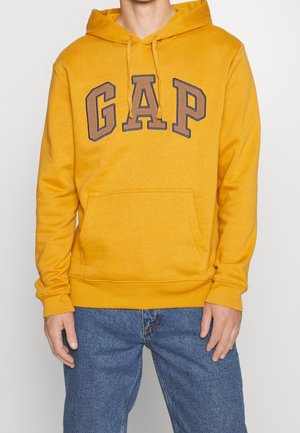 Mężczyzna w musztardowożółtej bluzie z kapturem z dużym logo „GAP” na piersi, w połączeniu z niebieskimi dżinsami, stoi z rozluźnionymi rękami.