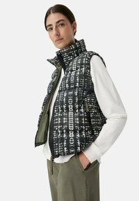 Vrouw in groene broek, wit langerhemd met lange mouwen en een zwart-wit patroon puffer vest, met haar handen in de zakken, kijkend naar de camera.
