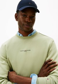 Felpa verde chiaro con collo rotondo, logo ricamato sul petto e una camicia blu piegata sotto. Cappellino blu scuro con logo.