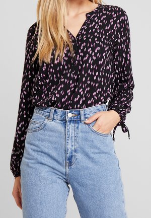 Femme portant un chemisier noir à manches longues avec un imprimé rose et blanc, rentré dans un jean en denim bleu clair taille haute, main dans la poche.