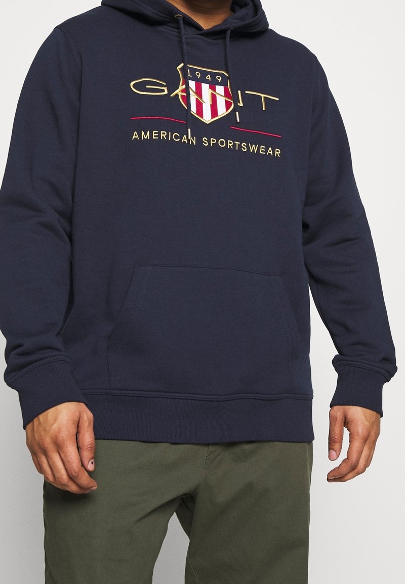 Sweat à capuche bleu marine avec poche kangourou avant, logo écusson brodé "GANT 1949" et texte "American Sportswear", porté avec un pantalon vert olive.
