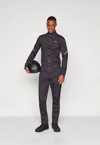 Vyriškas juodos spalvos dviračių kostiumas, sudarytas iš striukės su užtrauktuku ir atspindinčiomis detalėmis bei derančių kelnių, pagamintų iš glotnios, lengvos medžiagos. Laiko juodą šalmą.