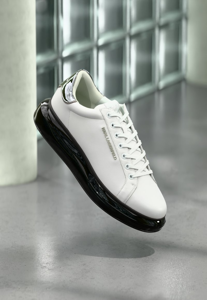 White leather sneaker with black rubber sole and transparent details. Features laces and embossed logo, set against a gray floor.