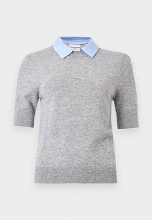 Frau trägt einen grauen Kurzarmpullover über einem hellblauen Hemd mit Kragen, einen langen dunkelblauen Rock und schwarze Ballerinas, steht vor weißem Hintergrund.
