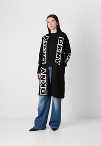 Črn kardigan z drznimi belimi logotipi "DKNY" ob sprednji strani in rokavih, v kombinaciji s širokimi modrimi traper hlačami in koničastimi črnimi čevlji.