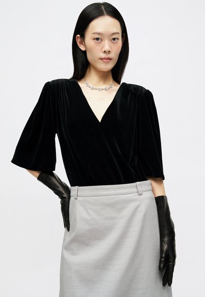 Γυναίκα με ίσια μαύρα μαλλιά, φορώντας μαύρη βελούδινη μπλούζα, ασημένιο κολιέ, γκρι φούστα και μακριά μαύρα δερμάτινα γάντια, ποζάρει μπροστά σε λευκό φόντο.