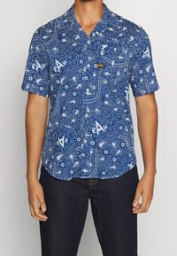 En person bär en kortärmad blå skjorta med vita mönster, som har olika former och en ficka. Mörka jeans fulländar outfiten.