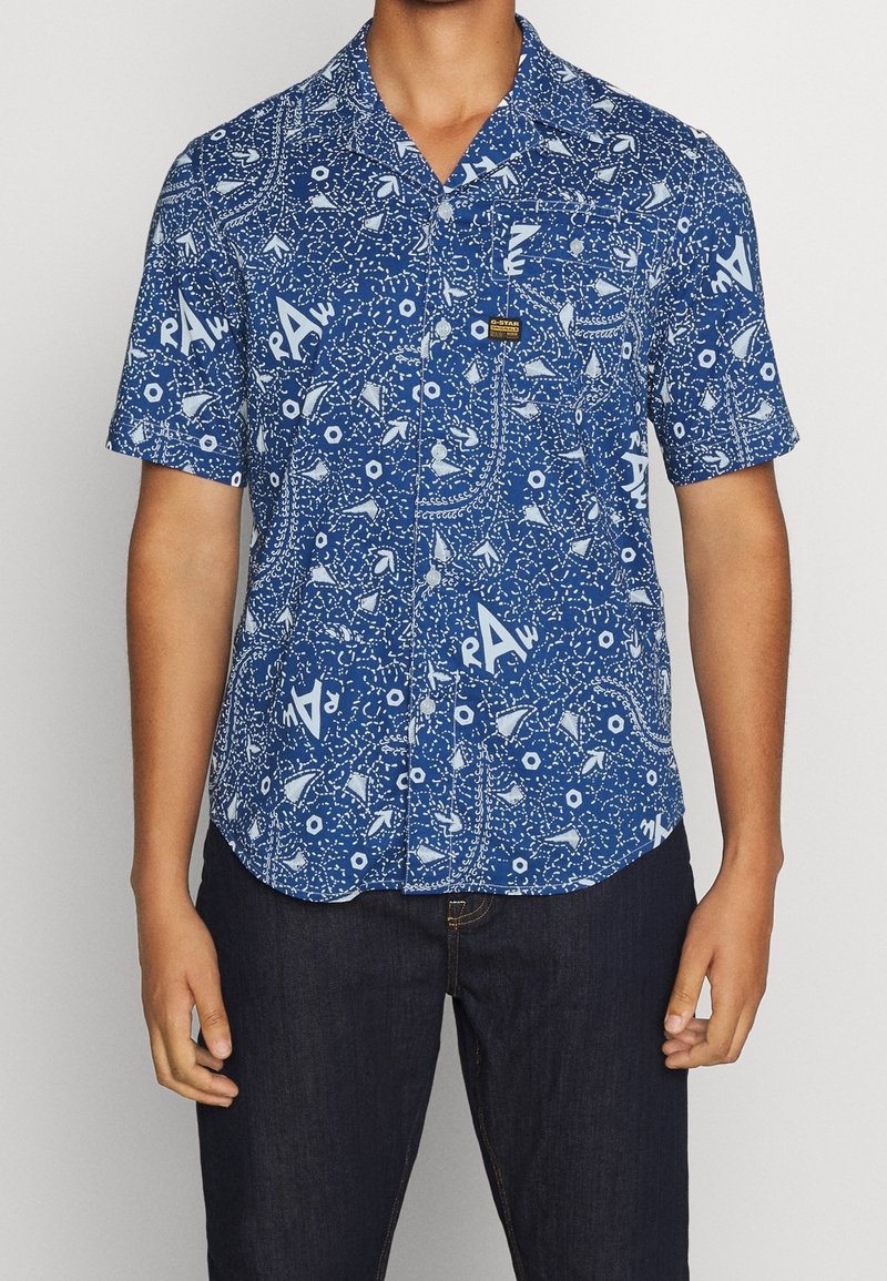 En person bär en kortärmad blå skjorta med vita mönster, som har olika former och en ficka. Mörka jeans fulländar outfiten.