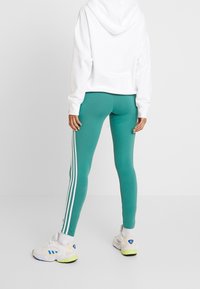 Person som bär tealleggings med vita sidostråk, vit hoodie och vita Adidas-sneakers med blå och gula detaljer, stående på ett vitt golv.