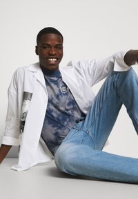 Camisa blanca de botones sobre una camiseta de tie-dye azul. Jeans de color azul claro con un diseño ajustado. El sujeto está sentado contra un fondo sencillo.