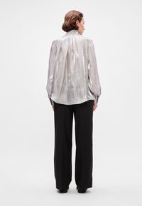 Silver, shiny blouse with a pleated design, high collar, and billowy sleeves, paired with black wide-leg trousers featuring small sparkles.