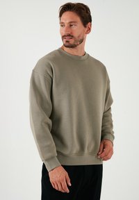 Sudadera verde oliva claro con cuello redondo, hombros caídos y puños y dobladillo de canalé. Textura suave, corte relajado.