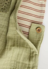 Ljust grönt tyg med en strukturerad yta, med ett randigt foder i beige och roströd samt en knappdetalj i trä på fickan.