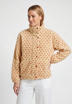 Kvinde iført beige fleecejakke med røde polkaprikker, stående med hånd på hoften iført hvide bukser mod en neutral baggrund.