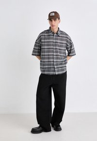Cămașă cu mânecă scurtă în carouri, neagră și gri, asortată cu pantaloni negri largi și pantofi negri. Pălăria este maro cu un patch cu logo.