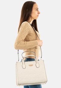 Bolso acolchado blanco con asas dobles y una correa para el hombro desmontable. Con detalles del logotipo en dorado. Textura suave, forma rectangular.