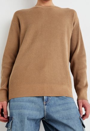 Personne portant un pull à manches longues côtelé marron et un pantalon cargo en denim bleu clair, debout devant un fond blanc uni.