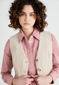 Gilet en velours côtelé beige avec deux boutons sur une chemise à carreaux rouge et blanc. Tissu texturé avec une coupe ajustée et un col.
