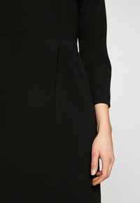 La main d'une femme repose à côté d'une robe noire à manches longues avec un détail de couture à la taille sur un fond clair.