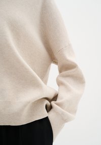 Pull en tricot beige avec des poignets et un ourlet côtelés. Le tissu semble doux avec une finition légèrement texturée. Associé à un pantalon noir.
