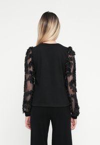 Top negro con mangas largas y transparentes adornadas con aplicaciones florales negras. La parte del cuerpo es completamente negra y tiene un escote redondo.