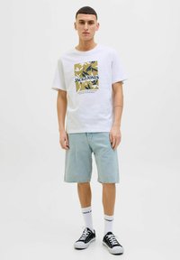 Hvid bomulds T-shirt med grafisk print i grøn og blå. Kombineret med lyse denimshorts og sorte sneakers med hvide accenter.