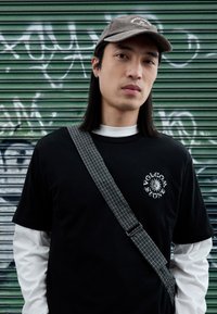 Μαύρο μπλουζάκι με άσπρο γραφικό σχέδιο, φορεμένο πάνω από λευκό μακρυμάνικο πουκάμισο. Γκρι καπέλο και τσάντα με καρό λουρί στον ώμο. Πράσινο φόντ ο.