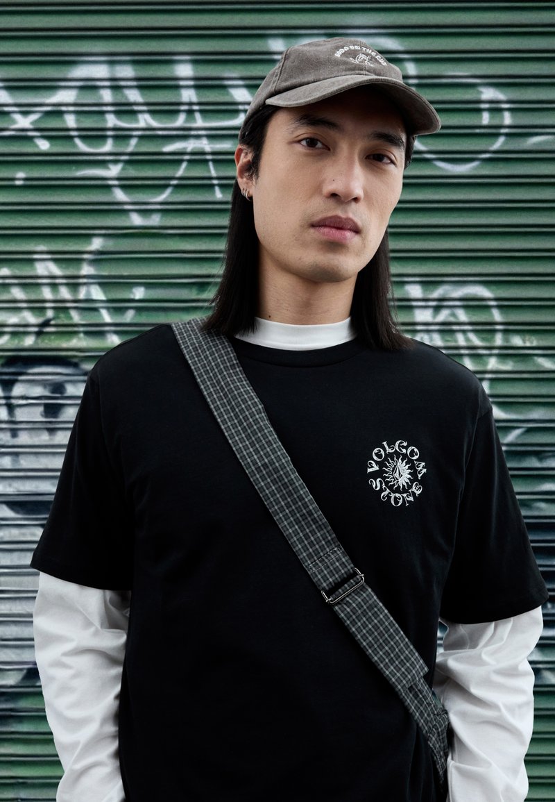 Μαύρο μπλουζάκι με άσπρο γραφικό σχέδιο, φορεμένο πάνω από λευκό μακρυμάνικο πουκάμισο. Γκρι καπέλο και τσάντα με καρό λουρί στον ώμο. Πράσινο φόντο.