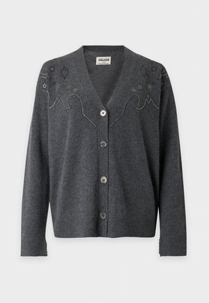 Une femme porte un cardigan brodé gris anthracite, un haut blanc, une jupe midi noire, des bottines et un sac à main noir à bandoulière chaîne, sur un fond uni.