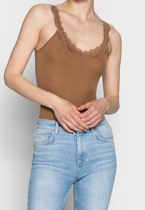 Vrouw draagt een mouwloos bruin topje met kanten rand en lichtblauwe high-waisted jeans, handen samengevouwen ter hoogte van de taille.