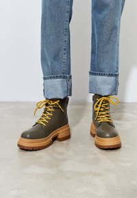 Botas de cuero verdes hasta el tobillo con cordones amarillos, suelas gruesas de plataforma beige y un acabado texturizado. Unos pantalones vaqueros con dobladillos terminados completan el look.