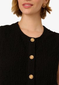 Zwarte textuur jas met een ronde halslijn en gouden knopen aan de voorkant. Gouden ronde oorbellen maken de look compleet.