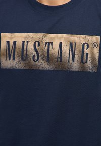 Nærbillede af en navyblå t-shirt med en gul rektangulær grafik og ordet "MUSTANG" i fede store bogstaver.