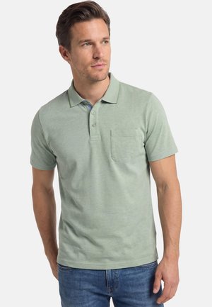 Mann trägt ein hellgrünes Polo-Shirt mit kurzen Ärmeln, Knöpfen am Kragen und Brusttasche, kombiniert mit blauen Jeans, blickt zur Seite.