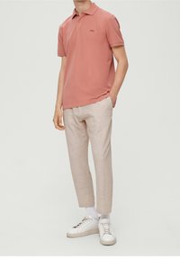 Man wearing a dusty pink short-sleeve polo shirt, light beige trousers, white socks, and white sneakers, standing with one hand in pocket.