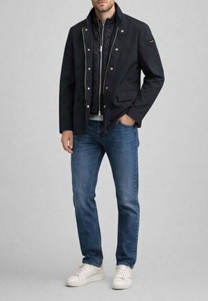 Uomo che indossa una giacca nera a strati con gilet interno trapuntato, jeans blu e scarpe da ginnastica bianche, in piedi con una mano in tasca.